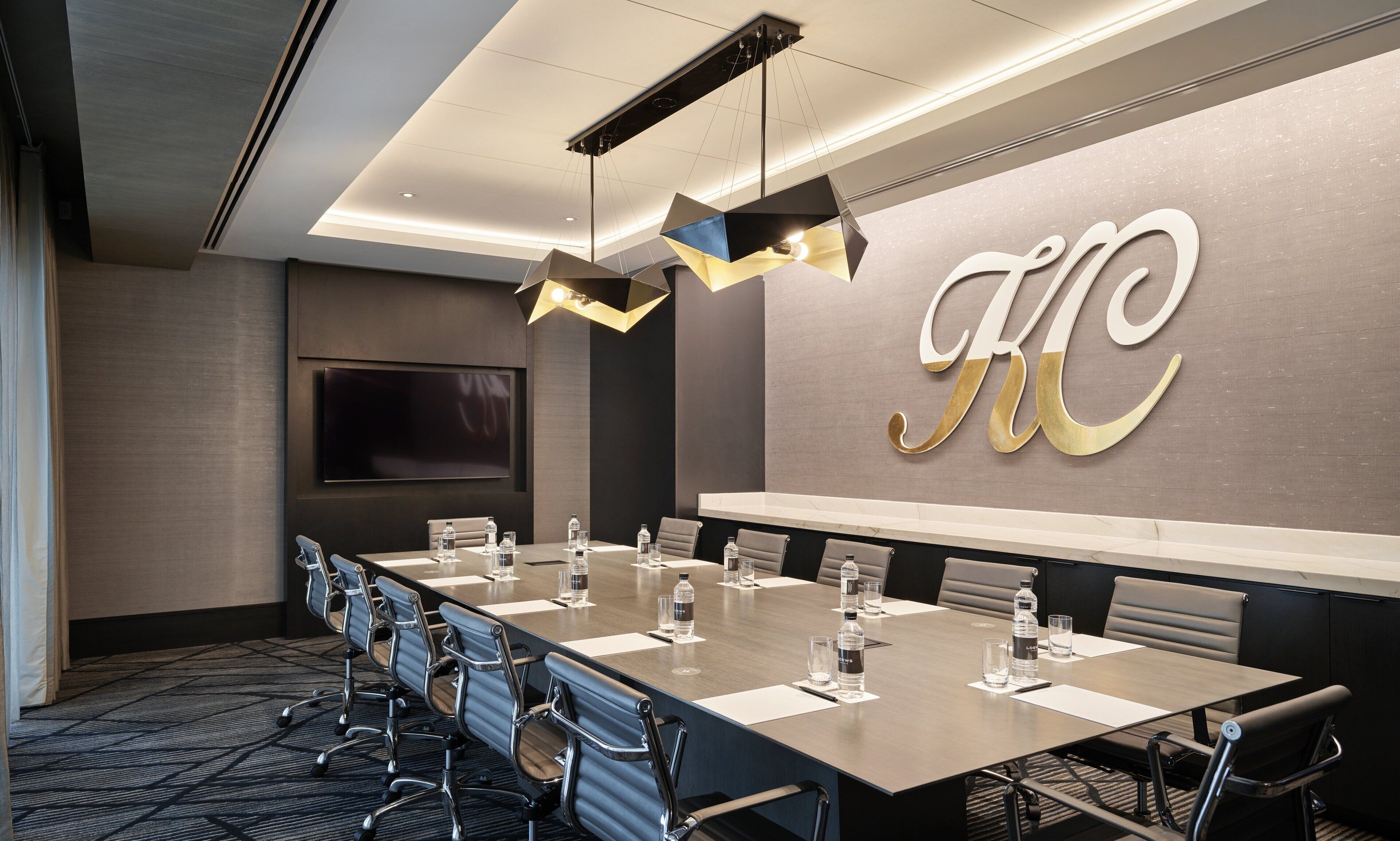 A conference room with a long table, chairs, a large screen, and a wall with a large letter.
