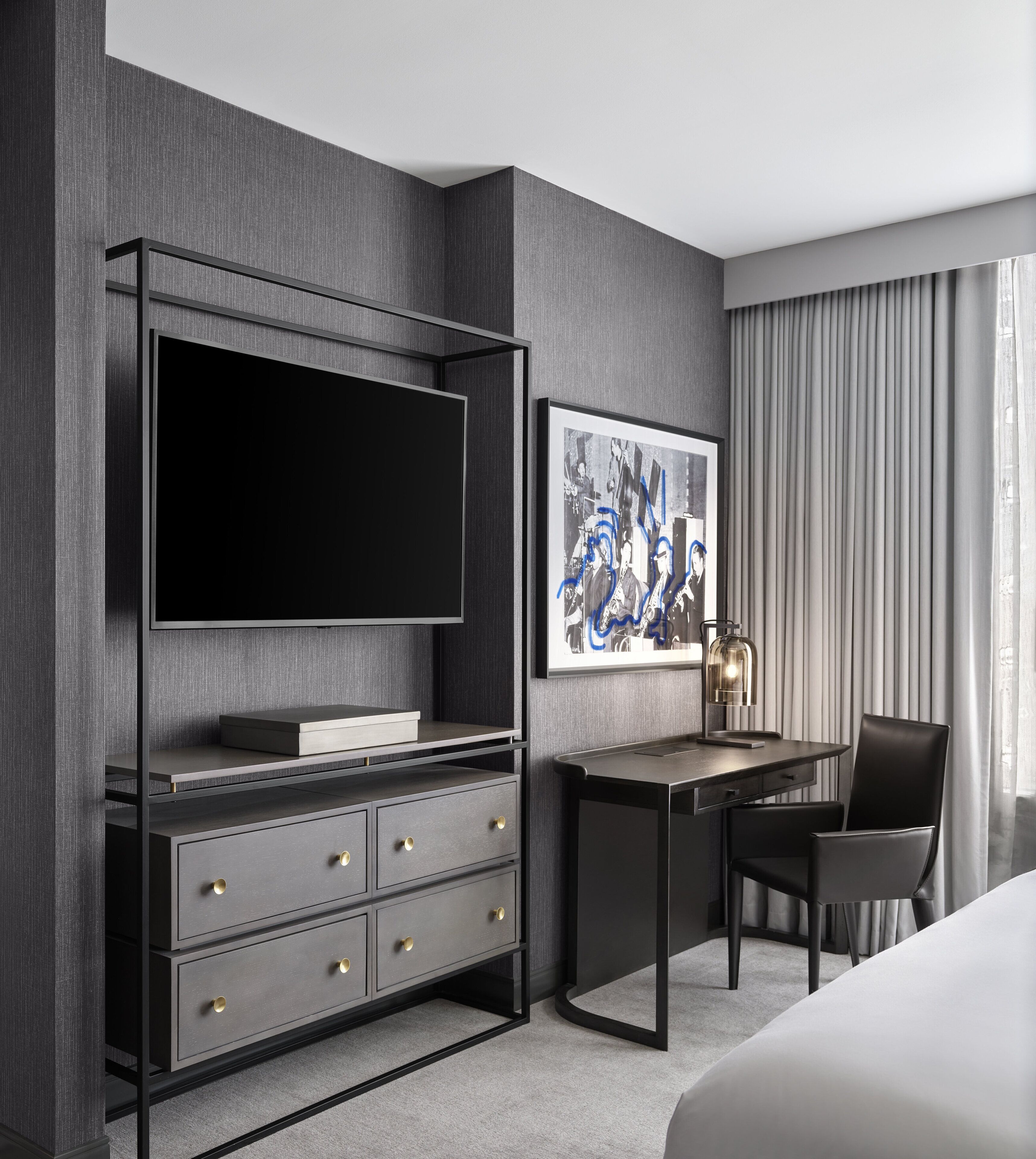 Une chambre d’hôtel moderne dotée d’une grande télévision à écran plat, d’un bureau avec une chaise, d’un lit et d’une œuvre d’art abstraite encadrée accrochée au mur.