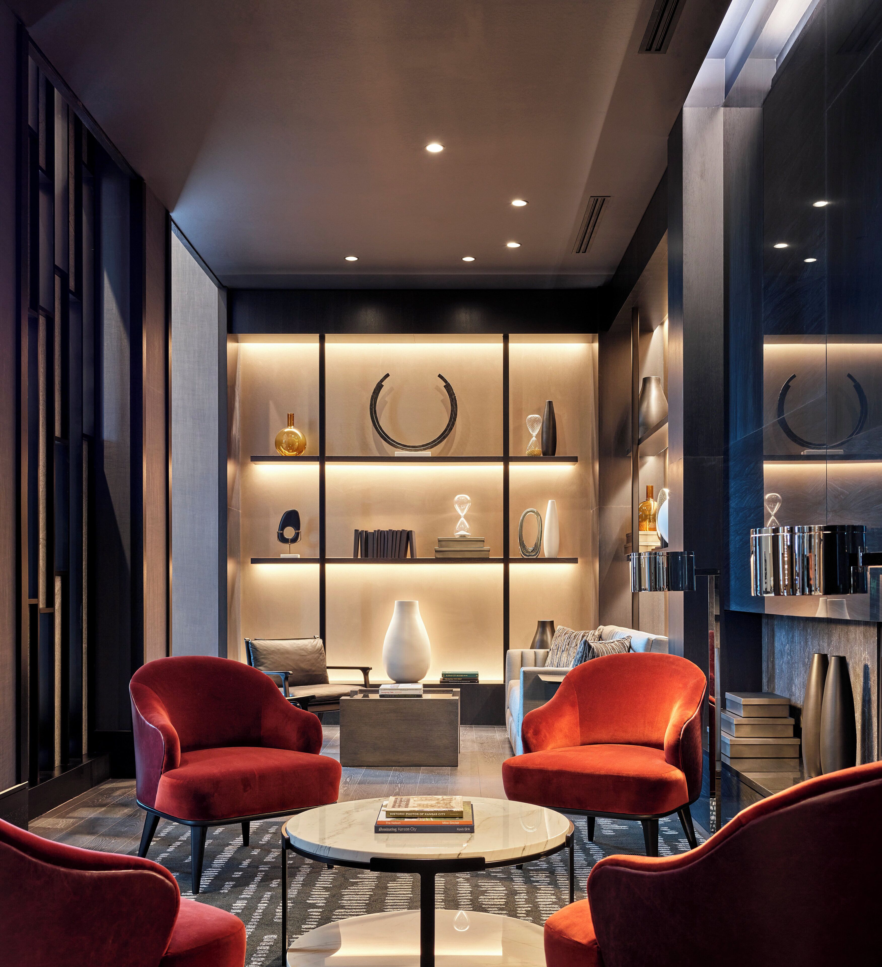 Un interior moderno con una zona de estar que incluye sillas tapizadas en rojo, una mesa de centro de mármol y una estantería con objetos decorativos.