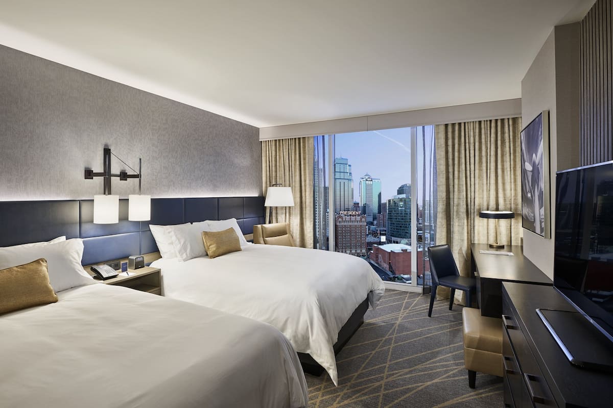 Habitación de hotel con dos camas, un escritorio, una silla, un televisor y vistas a la ciudad.