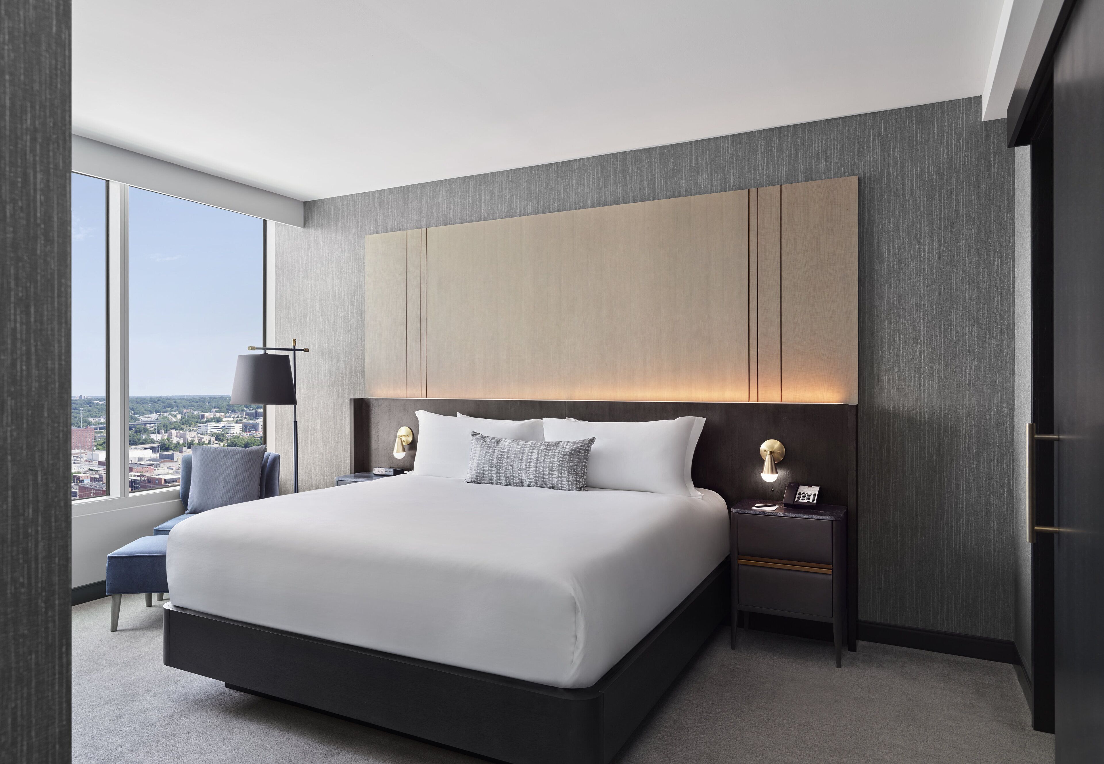 Een moderne hotelkamer met een groot bed, een nachtkastje, een lamp en uitzicht op de stad door grote ramen.