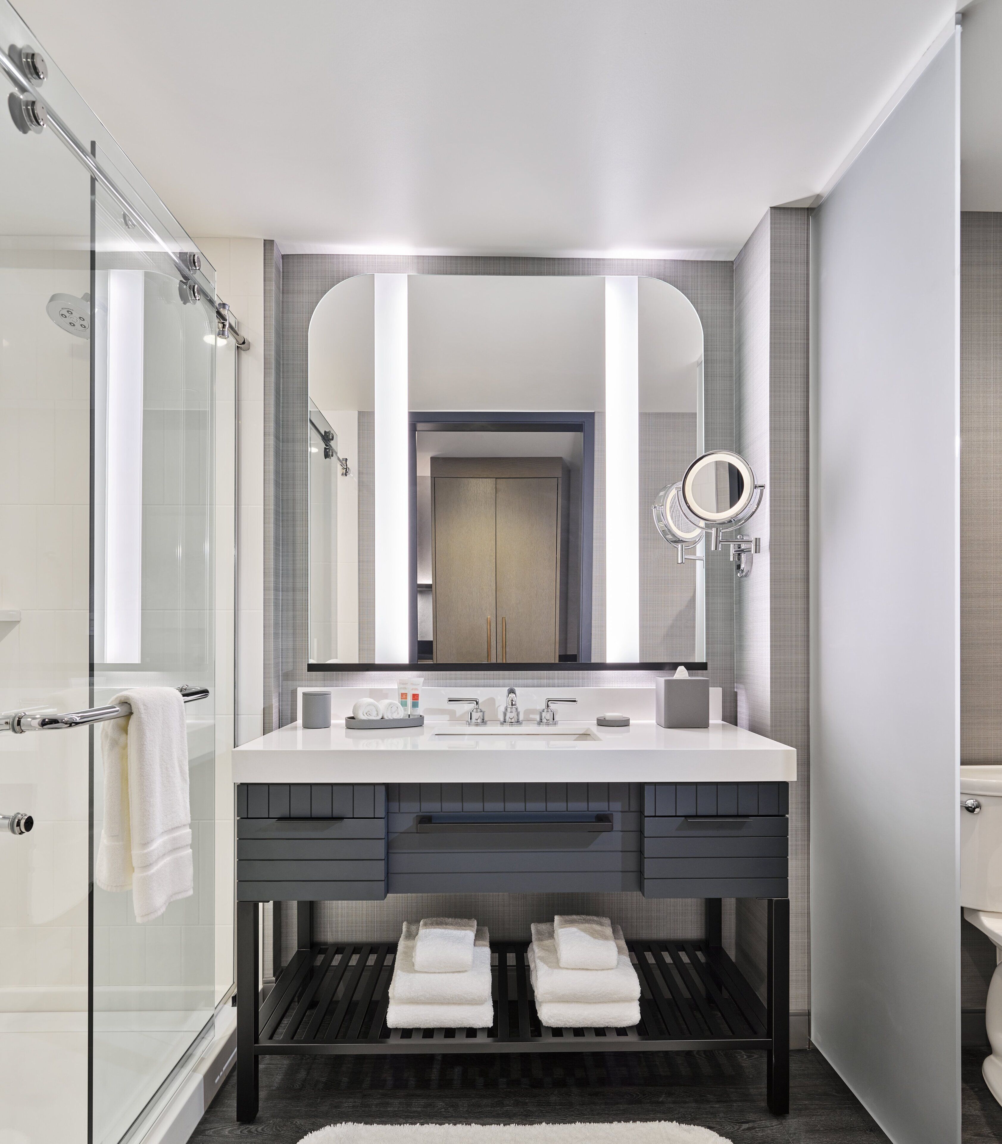 A modern bathroom with a glass shower enclosure, a large mirror, and a vanity with a dark countertop and drawers.