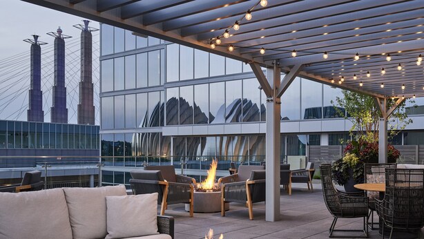 An outdoor seating area with a fire feature, modern furniture, and a view of a cityscape with buildings and a bridge. - Loews Kansas City Hotel (Kansas City)