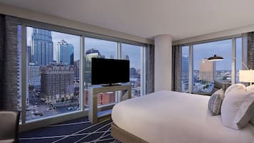 A hotel room with a large bed, a TV on a stand, and a city view through large windows.