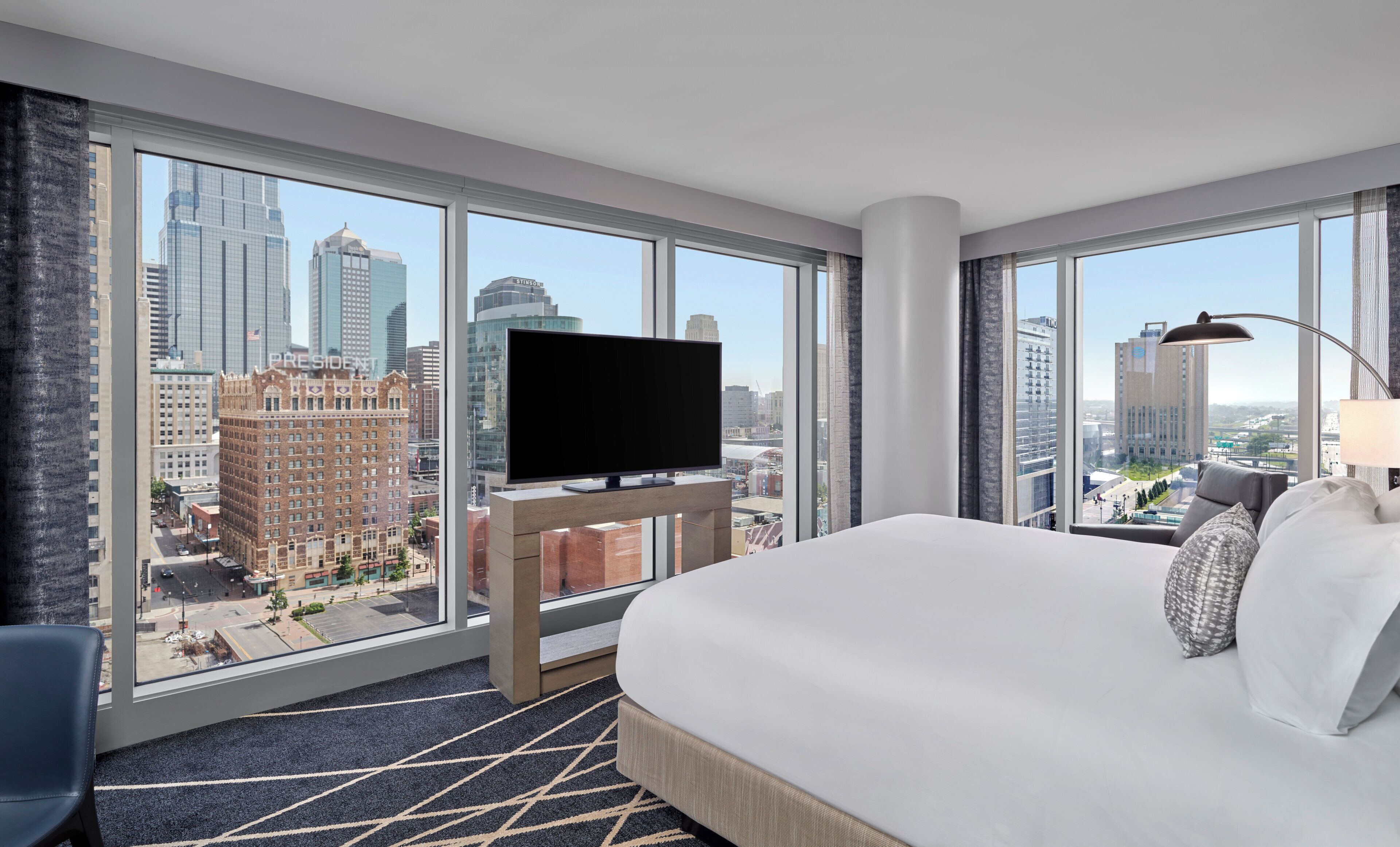 Une chambre d’hôtel dotée d’un grand lit, d’un téléviseur à écran plat posé sur un meuble, offrant une vue sur le paysage urbain grâce à de grandes fenêtres, et dont le sol est recouvert d’une moquette.