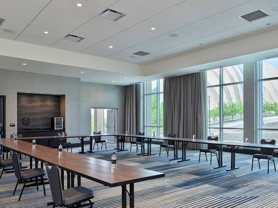 A conference room with a long table, chairs, a refrigerator, and large windows offering a view of a modern building and trees.