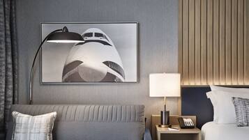 Una habitación de hotel con un sofá gris, una cama, una mesita de noche con una lámpara, un cuadro enmarcado de un avión y una pared de paneles de madera.