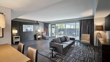 A modern hotel room with a sofa, dining table, and chairs, a TV, and a large window with a view of the outdoors.