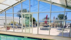Una piscina interior con paredes de vidrio transparente, suelo de baldosas y vistas a una zona de playa con sombrillas y sillas de descanso.
