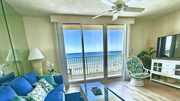 Una sala de estar con un sofá azul, una mesa de centro de vidrio y un sillón gris. Se observa un mueble para televisión con un televisor de pantalla plana y un ventilador de techo.