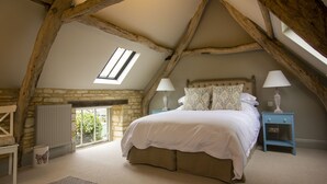 A bedroom with a sloped ceiling, stone walls, a large bed, two bedside tables with lamps, and a window. - Converted Luxury Barn - The Potting Shed - Grade II listed (Cirencester)
