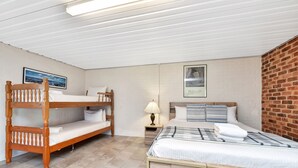 Une chambre avec des lits superposés et un lit simple, des murs blancs, un mur en briques apparentes et un tableau encadré accroché au mur.