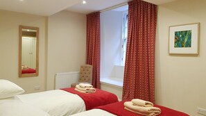 A hotel room with two beds, a large mirror, a radiator, a window with red curtains, and a framed picture on the wall.