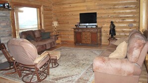 Ein rustikales Blockhaus-Interieur mit einem braunen Sofa, einem Schaukelstuhl, einem Fernseher auf einem Holzständer und einer Statue.