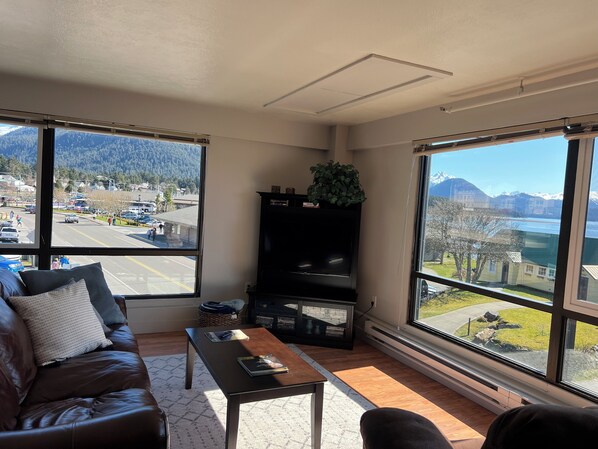 A living room with a leather couch, a coffee table, and a bookshelf. Large windows offer views of a parking lot and mountains. - Downtown Apt 6 w/ Panoramic Ocean & Mountain Views (Sitka)
