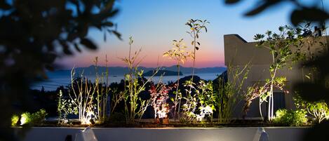 Una terraza con dos sillas de plástico blancas y una mesa redonda, con vista a un paisaje escénico del océano y las montañas al atardecer.