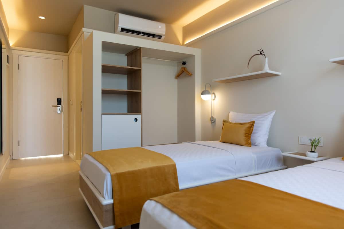 Una habitación de hotel con dos camas, un armario, una mesita de noche con lámpara y una estantería en la pared.