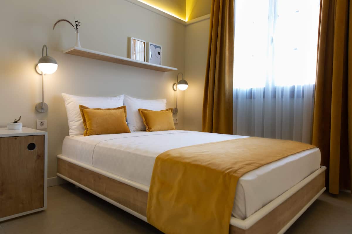Una cama bien hecha con ropa de cama blanca y amarilla, dos lámparas de noche, una mesita auxiliar pequeña y un estante sobre la cama.