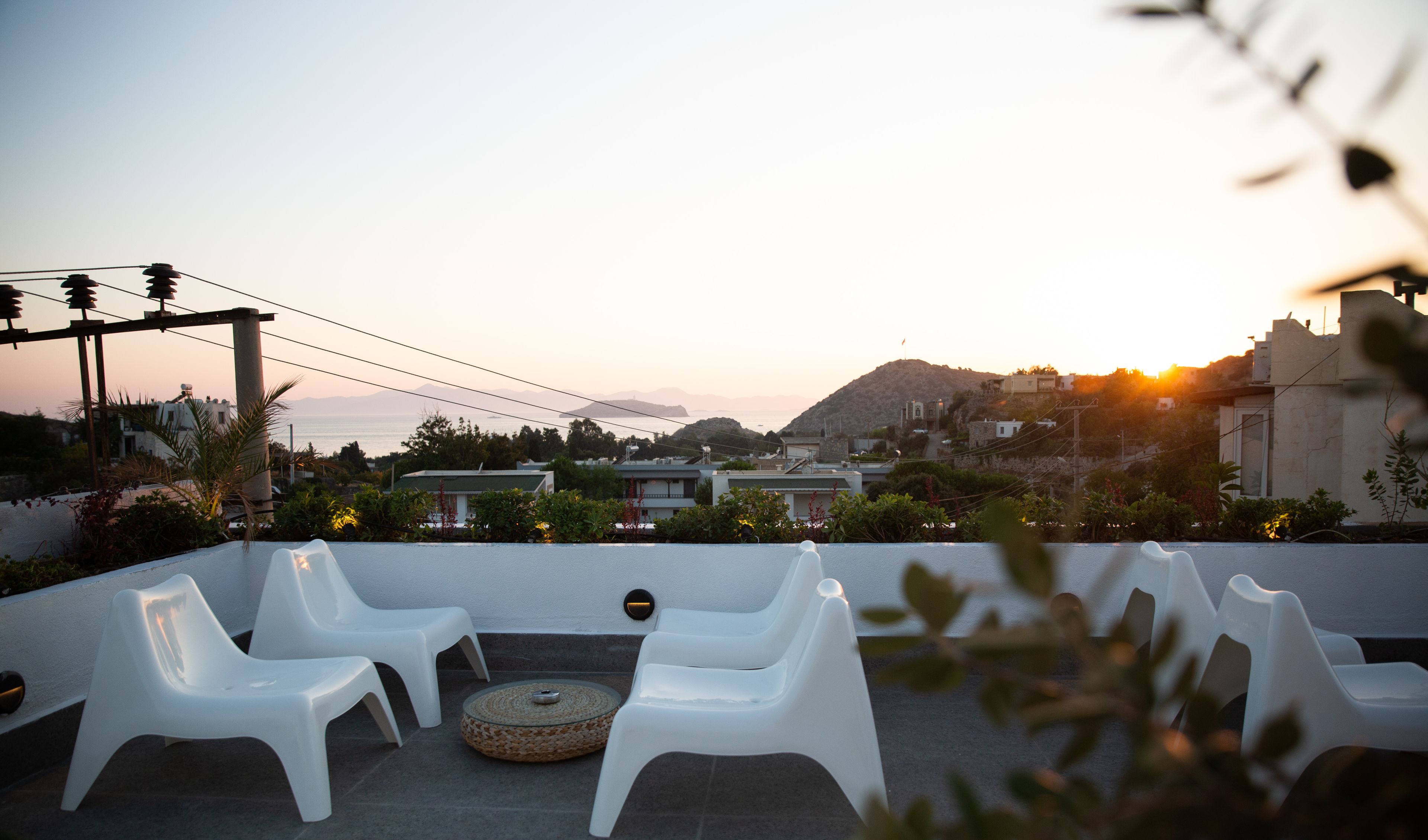 A rooftop terrace with white plastic chairs and a woven table, overlooking a coastal town with buildings and a mountain in the background.