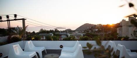 A rooftop terrace with white plastic chairs and a woven table, overlooking a coastal town with buildings and a mountain in the background.