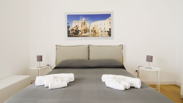 Una cama bien hecha con ropa de cama gris, dos almohadas y toallas blancas dobladas sobre la cama. Hay dos mesitas de noche con lámparas y una fotografía enmarcada de un edificio histórico en la pared.