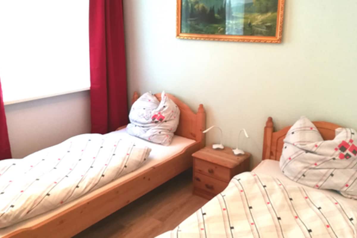 Un dormitorio con dos camas individuales, una mesita de noche y una ventana con cortinas rojas.