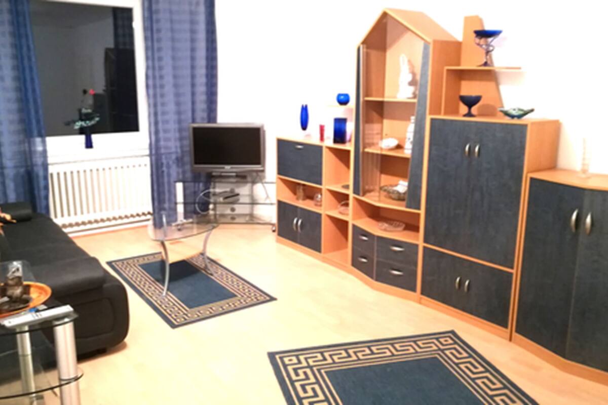Una sala de estar con un sofá negro, una mesa de centro de vidrio, un televisor y armarios de madera con estantes.