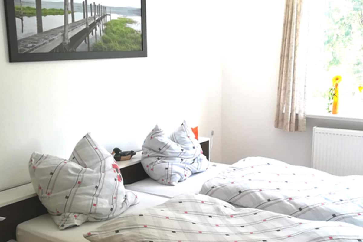 Una cama individual con una colcha blanca y roja de estampado, una mesita de noche y un cuadro enmarcado con una imagen de un muelle.