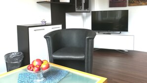 Un salon avec un fauteuil en cuir noir, une table en verre sur laquelle sont posées des pommes, un téléviseur et une étagère ornée d’objets décoratifs.