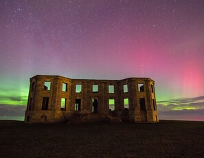 Un bâtiment historique doté de nombreuses fenêtres et d’une porte d’entrée centrale, se dressant sur fond de ciel nocturne parsemé d’étoiles et illuminé d’une aurore boréale verdoyante.