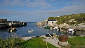 A coastal scene with boats docked at a stone pier, a grassy hill, and a small building. - Porthole Cottage, Bushmills -
 a place to unwind and relax. (Dunseverick)
