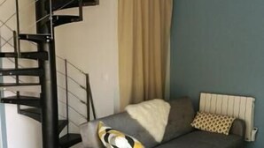 Un salon moderne avec un canapé gris, un tapis blanc et moelleux, une table basse en verre, un rideau beige et un escalier en colimaçon noir.