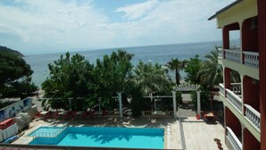 Zona de piscina con vista al mar, rodeada de palmeras y sillas rojas.