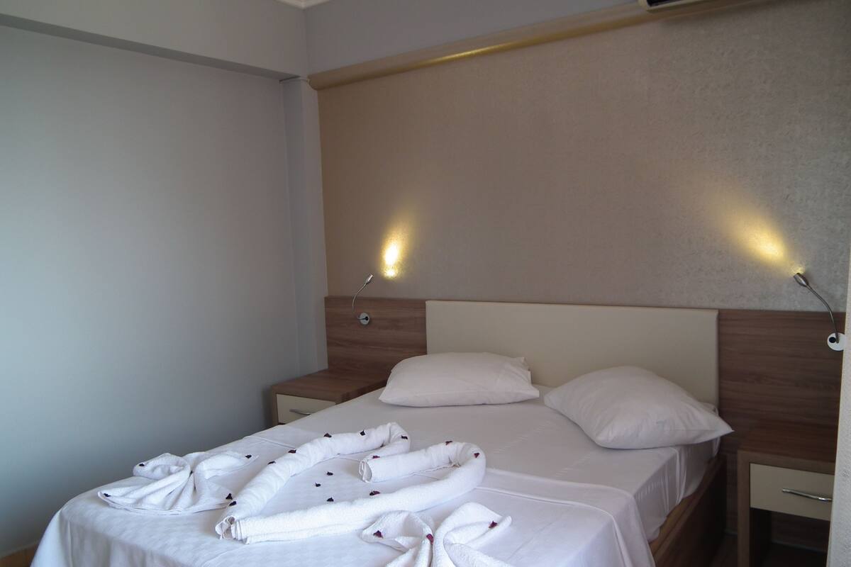 A hotel room with a bed, two bedside tables, a wall-mounted air conditioner, and two wall-mounted lights.
