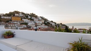Una terraza con una zona de estar blanca acolchada, plantas en macetas y una botella de vino en una barandilla, con vistas a un pueblo costero de edificios blancos y al mar.