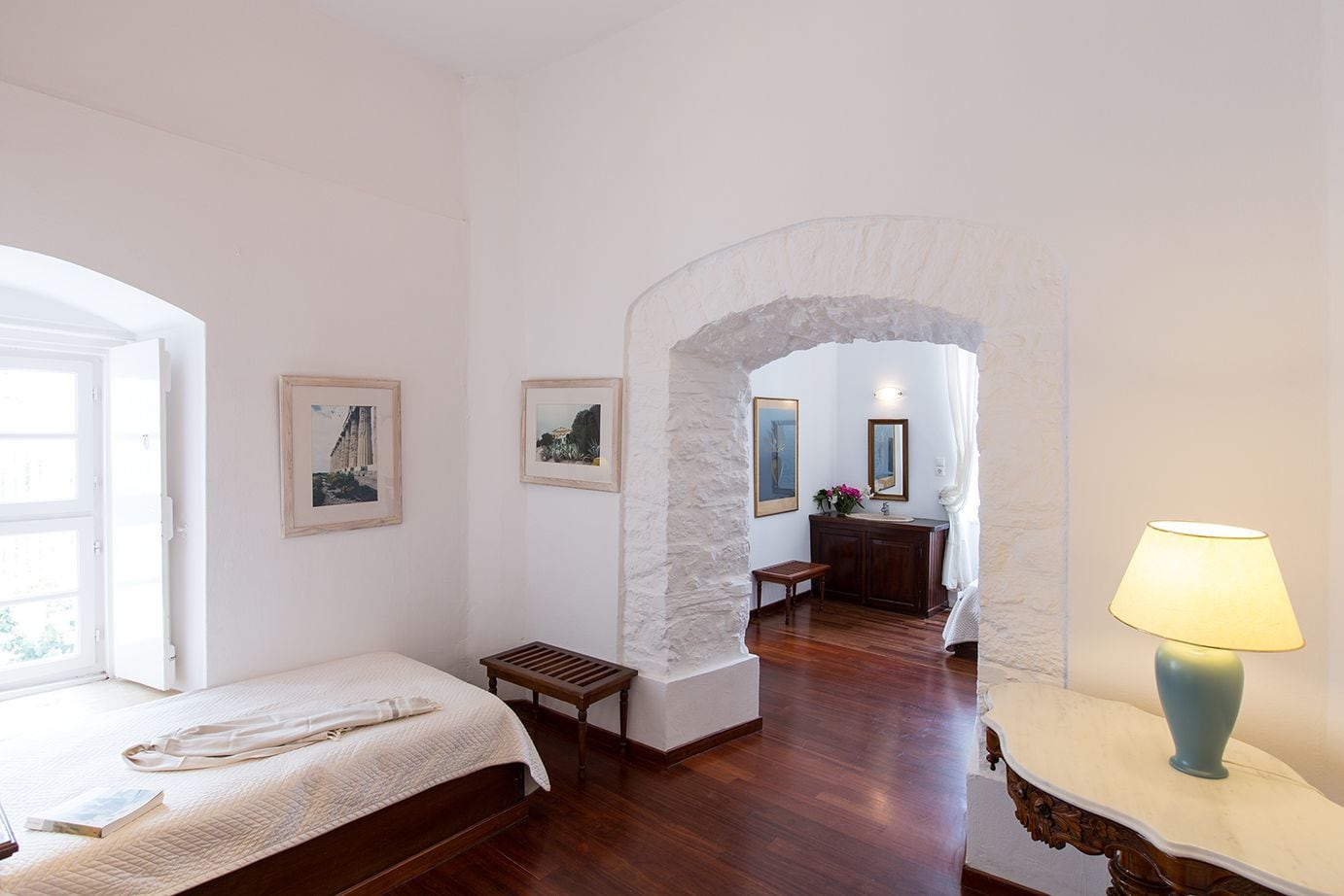 Une chambre à coucher comprenant un lit, un banc, une table de chevet et une lampe. Une porte donne accès à une salle de bain équipée d’un lavabo et d’un miroir.