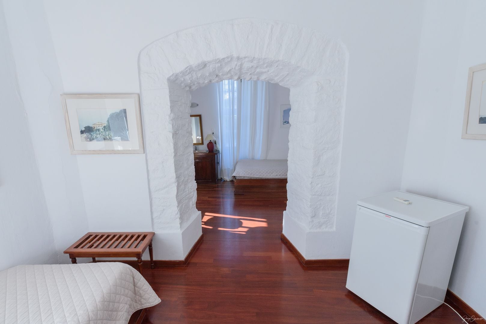 A room with a wooden bench, a bed, a white refrigerator, and a white wall with a framed picture.