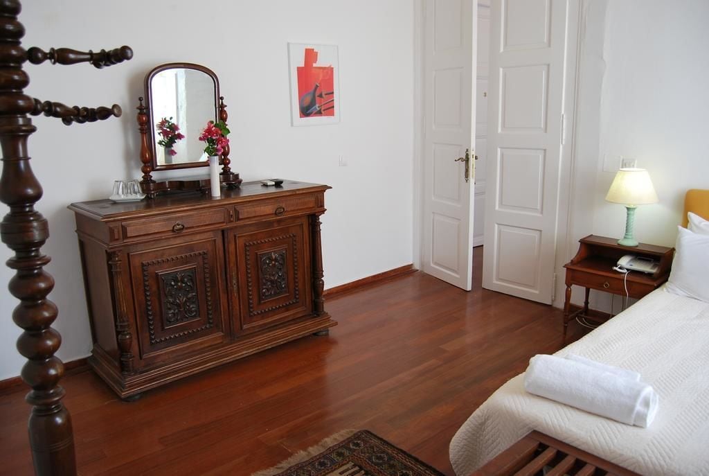 Un dormitorio con un aparador de madera, una cama con edredón blanco, una mesita de noche con lámpara, y una puerta que da a otra habitación.
