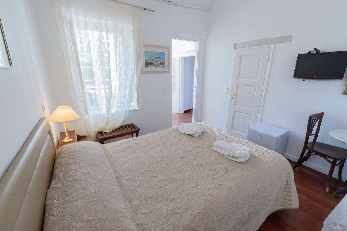 Habitación de hotel con una cama grande, una mesita de noche con lámpara, una silla, un escritorio con computadora y televisor en la pared.