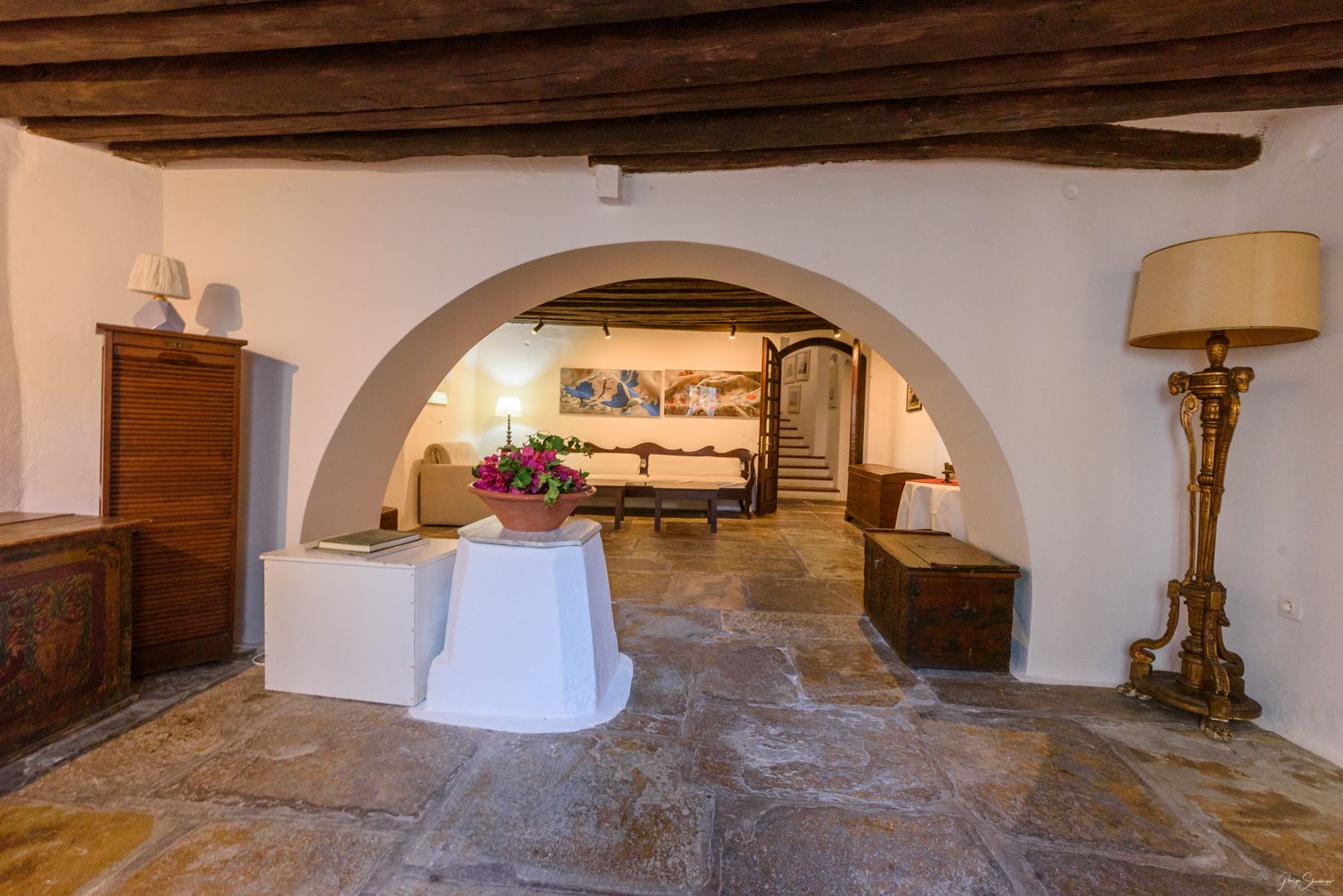 A stone-floored hallway with an arched doorway leading to a bedroom. A white table with a flower pot is placed in the center.