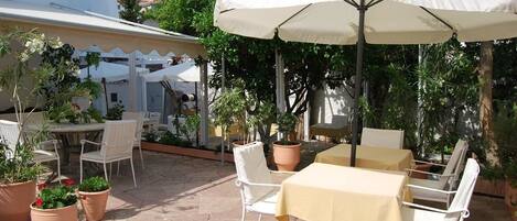 Une terrasse extérieure avec des chaises et des tables blanches, protégée par un grand parasol et entourée de plantes en pot.