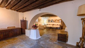 Un interior rústico con suelo de piedra, mostrador de recepción de madera, un pedestal blanco con maceta, un sofá y una escalera que sube a un nivel superior.