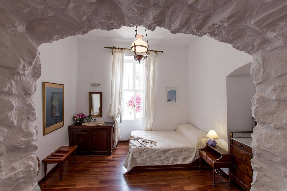 Un dormitorio con un arco de piedra, suelo de madera, una cama con cubrecama blanco, una mesita de noche con lámpara, un aparador, un tocador con espejo y un cuadro enmarcado en la pared.