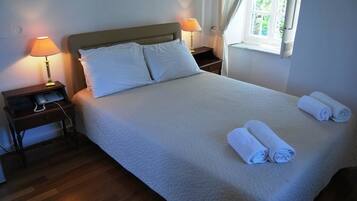 A neatly made bed with white linens and pillows, two bedside lamps, a wooden nightstand, and a window with curtains.