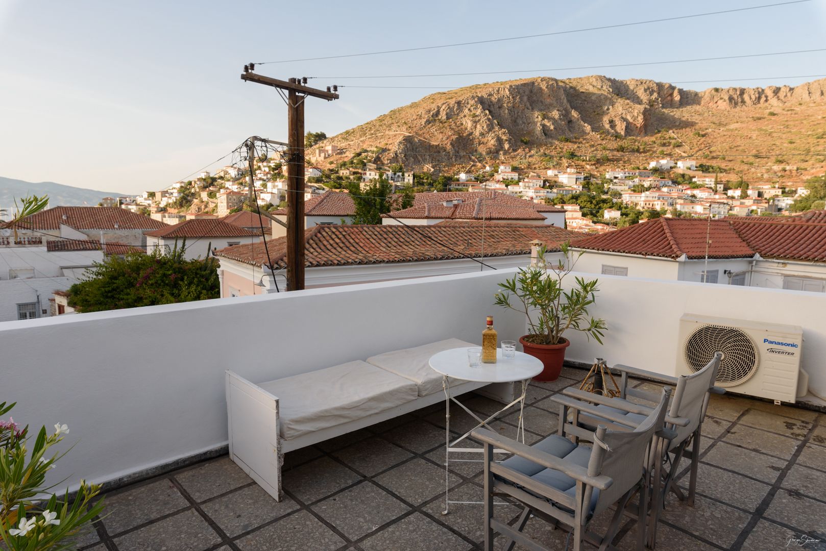 A rooftop terrace with a white sofa, a small round table, and two chairs. There is a potted plant and a Panasonic air conditioner. The terrace overlooks a town with red-tiled roofs and a mountain in the background.