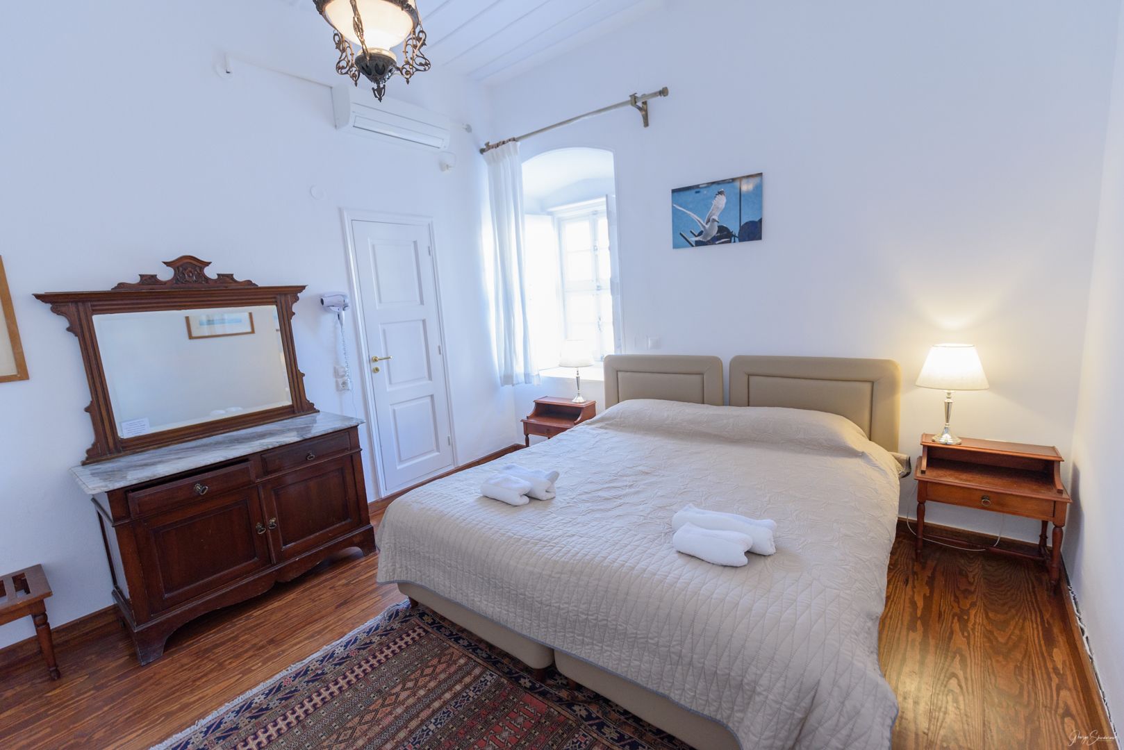 A bedroom with a large bed, a wooden dresser, a mirror, a nightstand with a lamp, and a window with curtains.