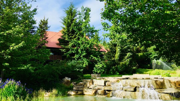A tranquil garden with a small waterfall, lush greenery, and a building with a red roof. - Amazing location steps from Whistler Village, with outdoor heated pool/hot tub! (Whistler)