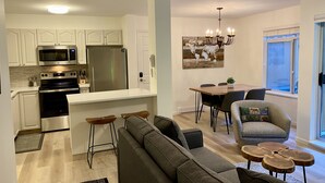 A modern kitchen with white cabinets, a stainless steel refrigerator, and a built-in microwave. There is a dining area with a wooden table and chairs, and a gray sofa with a small coffee table. - Amazing location steps from Whistler Village, with outdoor heated pool/hot tub! (Whistler)