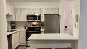 A modern kitchen with white cabinetry, a stainless steel refrigerator, a microwave, and an oven. There are two wooden stools at the kitchen island. - Amazing location steps from Whistler Village, with outdoor heated pool/hot tub! (Whistler)