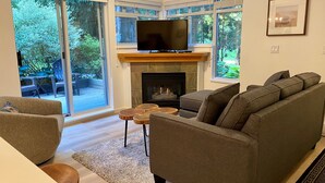 A living room with a fireplace, a sofa, a TV, and large windows overlooking a garden. - Amazing location steps from Whistler Village, with outdoor heated pool/hot tub! (Whistler)